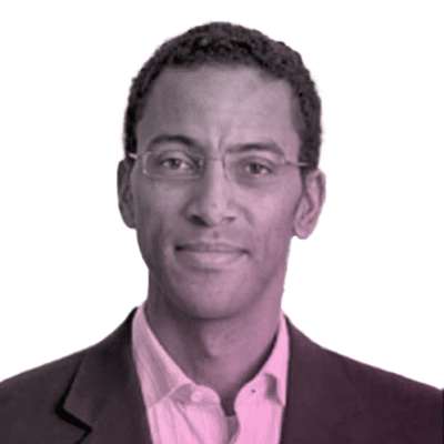 A man with short curly hair and glasses wearing a suit jacket and collared shirt looks at the camera against a white background.