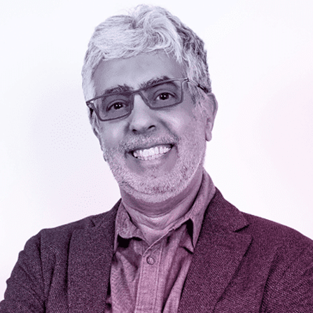 A man with short gray hair and glasses, wearing a blazer over a button-up shirt, smiles at the camera against a plain background.