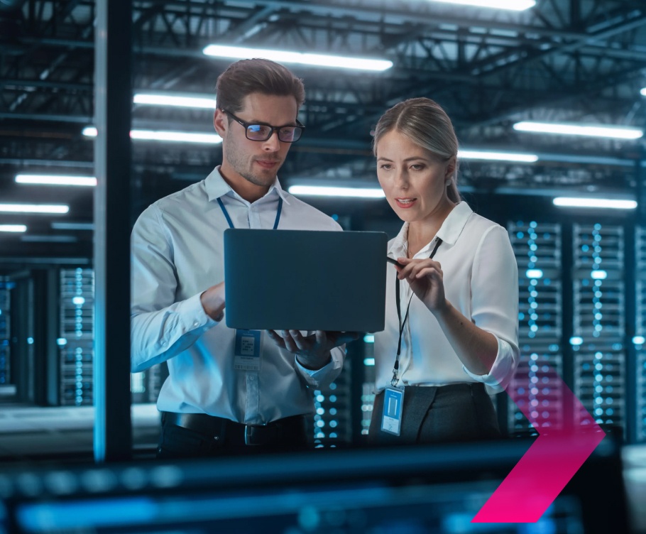 Two professionals from security teams stand in a server room, looking at a laptop together. They are both wearing ID badges and discussing important information displayed on the screen.
